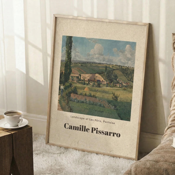 May include: Framed art print of rural countryside landscape with rolling green hills, farmhouse buildings, and cloudy blue sky, displayed on white carpeted floor against white wall corner. Warm wooden frame surrounds cream-colored matting. Small wooden stool with white coffee cup and books visible to left. Soft natural lighting creates gentle shadows across the scene.