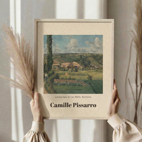 May include: Person in cream ruffled sleeves holding framed vintage landscape print showing rural French countryside with rolling hills, farm buildings, and cloudy sky. Dried pampas grass visible to left. Artwork features muted greens, blues, and earth tones in impressionist style within light wooden frame against soft neutral background.