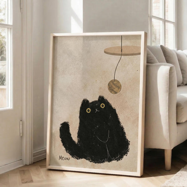 May include: Large framed poster leaning against white wall next to beige sofa in bright living room. The artwork shows a black textured cat with golden eyes sitting upright, looking up at a hanging ball of yarn. The cat appears painted in rough, fuzzy brushstrokes with "Meow" text below. The frame is light wood against cream-colored paper background. Natural sunlight streams through tall windows.