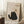 May include: Framed poster artwork featuring a black fuzzy cat with yellow eyes sitting upright with the word "Meow" below. Above the cat hangs a brown yarn ball from a curved line. The artwork has a cream textured background in a light wood frame, displayed on a white shag rug next to a wooden side table with coffee cup and saucer. Soft natural lighting creates window shadows on white walls.