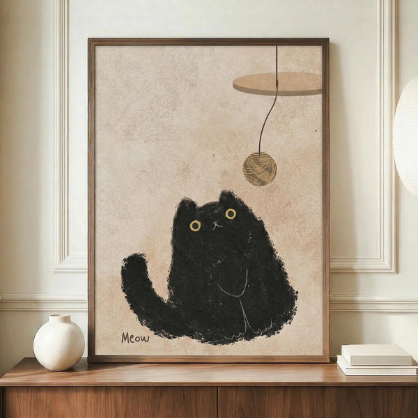 May include: Framed artwork displayed on wooden sideboard showing a fluffy black cat with yellow eyes reaching up toward a dangling ball of yarn, with "Meow" text below. The illustration has a textured, sketchy style on beige background. Scene includes white ceramic vase and stacked books as decor against cream walls with white trim molding.