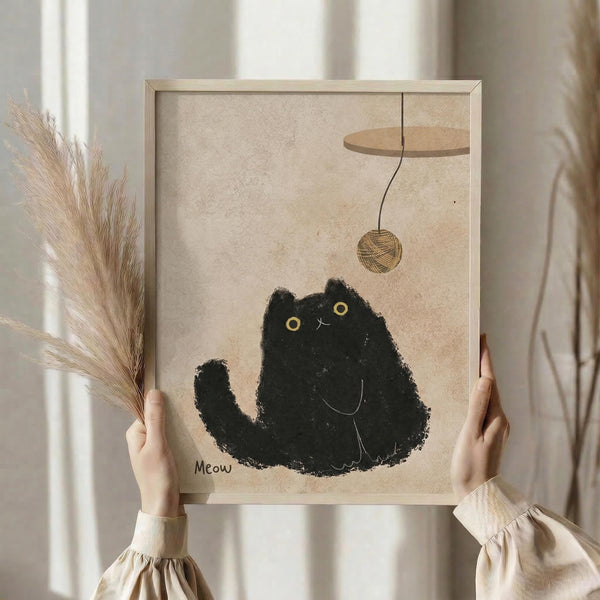 May include: Person holding framed artwork showing black cat silhouette with yellow eyes playing with brown yarn ball, with "Meow" text below. Warm beige textured background, natural wood frame. Soft neutral tones throughout scene with dried pampas grass visible on left side. Cozy minimalist home decor styling.