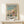 May include: Framed poster print featuring an impressionist beach scene with people in period clothing on sandy shore, turquoise water and cliffs in background. Poster shows soft brushstrokes in blues, pinks, and yellows with Claude Monet signature at bottom. Natural wood frame sits on light wooden surface beside small wrapped gift with twine, displayed against cream colored wall.