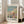 May include: Framed art print featuring Claude Monet's impressionist beach scene with figures in period dress enjoying a sunny coastal day, displaying soft blues, greens, and coral tones. The print shows Monet's signature and an inspirational quote, mounted in a light wooden frame leaning against a beige sofa in a bright, minimalist living room with white walls and natural lighting from tall windows.