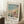 May include: Framed art print leaning against white wall on fluffy white rug next to wooden side table with teacup and beige throw blanket. Print shows impressionist beach scene with people in period dress holding colorful parasols on sandy coastline with turquoise water. Features Claude Monet signature and quote below artwork in cream matted frame.