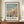 May include: Framed art print featuring an Impressionist beach scene with figures in Victorian dress holding parasols on sandy coastline, displayed on wooden sideboard beside white ceramic vase and books against cream wainscoted wall. Print shows soft brushstrokes in blues, greens, and coral tones with artist signature and quote below the image.