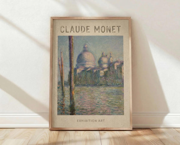 May include: Framed art poster featuring Claude Monet's impressionist painting of Venice's San Giorgio Maggiore church with distinctive domes across water, shown in soft blues, greens, and pinks. The poster has beige background with "CLAUDE MONET" and "EXHIBITION ART" text, displayed in light wood frame leaning against white wall on wooden floor with natural lighting casting gentle shadows.