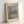 May include: Framed art print featuring Claude Monet's impressionist painting of San Giorgio Maggiore church in Venice, showing the domed basilica across shimmering water with wooden posts in foreground. Soft pastel colors in blues, pinks and yellows capture reflected light on water. Natural wood frame with cream matting displays "CLAUDE MONET" and "EXHIBITION ART" text. Print leans against white wall corner.