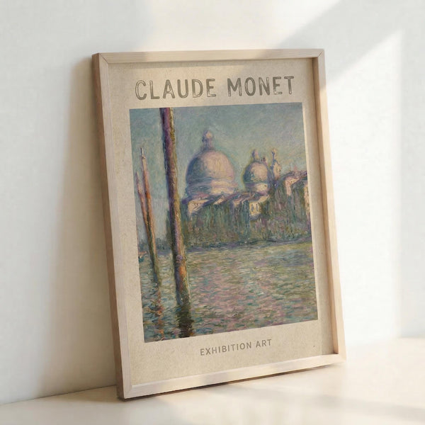May include: Framed art print featuring Claude Monet's impressionist painting of San Giorgio Maggiore church in Venice, showing the domed basilica across shimmering water with wooden posts in foreground. Soft pastel colors in blues, pinks and yellows capture reflected light on water. Natural wood frame with cream matting displays "CLAUDE MONET" and "EXHIBITION ART" text. Print leans against white wall corner.