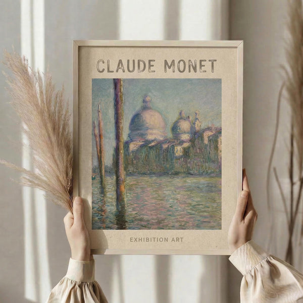 May include: Hands holding a framed vintage-style poster featuring a Monet impressionist painting of Venice's domed cathedral across shimmering water with vertical posts. The beige matted frame is displayed against soft cream curtains with dried pampas grass visible to the left. The artwork shows soft blues, greens and warm tones in Monet's characteristic brushstroke style, with "CLAUDE MONET" and "EXHIBITION ART" text above and below the image.