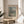 May include: Framed exhibition poster featuring Claude Monet's impressionist painting of Venice's San Giorgio Maggiore church with its distinctive domes reflected in water, displayed on a light wood round side table next to a beige upholstered chair with white throw pillow, green plant leaves visible, soft natural lighting creating shadows on cream wall
