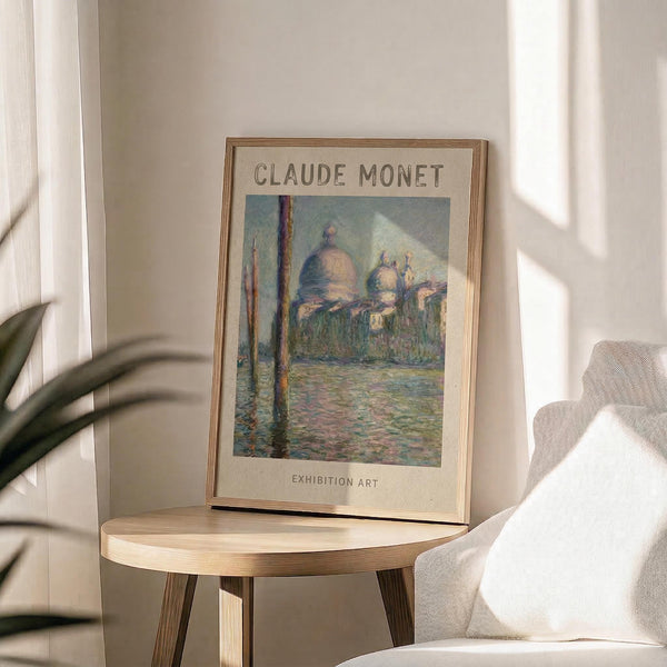 May include: Framed exhibition poster featuring Claude Monet's impressionist painting of Venice's San Giorgio Maggiore church with its distinctive domes reflected in water, displayed on a light wood round side table next to a beige upholstered chair with white throw pillow, green plant leaves visible, soft natural lighting creating shadows on cream wall