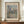 May include: Framed Claude Monet exhibition poster displaying a soft impressionist painting of Venice's San Giorgio Maggiore church with domed architecture reflected in shimmering water, featuring muted blues, pinks and greens. The beige matted print sits on a wooden console table beside a white ceramic vase and stack of books against cream paneled walls.