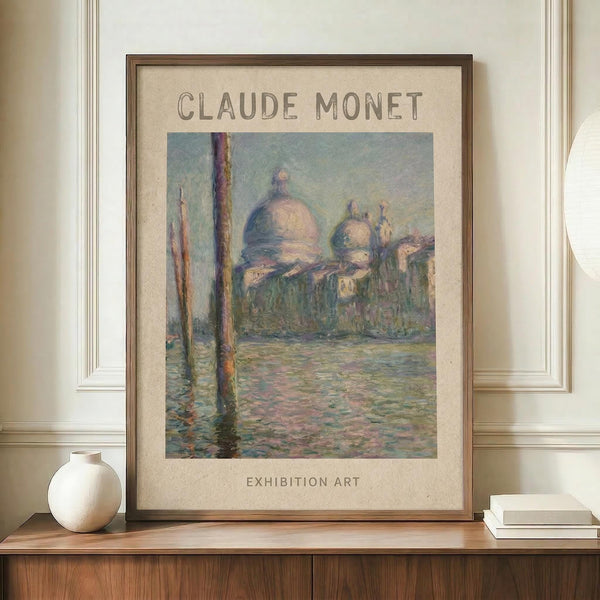 May include: Framed Claude Monet exhibition poster displaying a soft impressionist painting of Venice's San Giorgio Maggiore church with domed architecture reflected in shimmering water, featuring muted blues, pinks and greens. The beige matted print sits on a wooden console table beside a white ceramic vase and stack of books against cream paneled walls.
