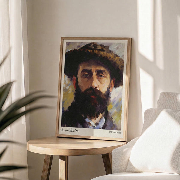 May include: Framed art print of a bearded man wearing a dark hat, displayed on a round wooden side table next to a beige upholstered chair. Natural sunlight streams across the white wall, creating soft shadows. The portrait shows expressive brushwork in warm earth tones with visible paint texture. A green plant leaf appears in the corner, adding to the cozy interior setting.