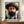 May include: Framed poster of a bearded man in a wide-brimmed hat, painted in bold brushstrokes with vibrant blues, greens, and earth tones. The impressionist-style portrait shows expressive eyes and thick facial hair against a colorful background. The artwork sits on a wooden sideboard next to a white ceramic vase and books in a bright room with cream walls and white trim.