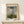 May include: Framed reproduction of impressionist painting showing woman in white dress holding green parasol on grassy hill with child figure, displayed on light wood mantel beside small wrapped white gift with twine and evergreen sprig against cream wall