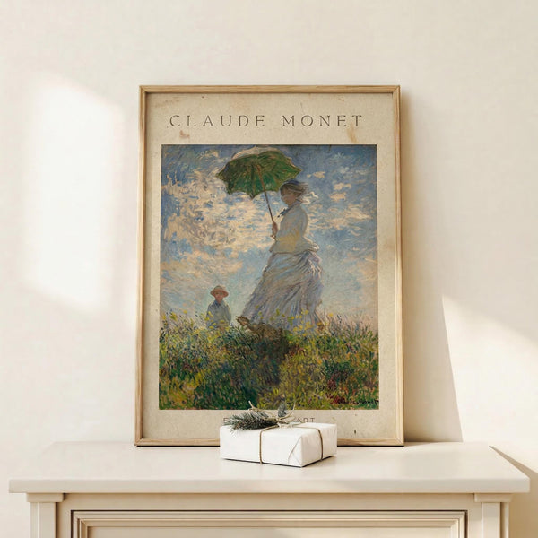 May include: Framed reproduction of impressionist painting showing woman in white dress holding green parasol on grassy hill with child figure, displayed on light wood mantel beside small wrapped white gift with twine and evergreen sprig against cream wall