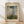May include: Framed art print featuring Monet's impressionist painting of a woman in a flowing white dress holding a green parasol, with a small child visible in tall grass below. The artwork shows soft blues and whites in the cloudy sky with vibrant greens in the foreground. The poster has aged cream-colored matting with "CLAUDE MONET" at top and "EXHIBITION ART" at bottom, displayed in a thin wooden frame leaning against white wall on light wood flooring.