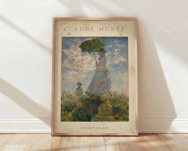 May include: Framed art print featuring Monet's impressionist painting of a woman in a flowing white dress holding a green parasol, with a small child visible in tall grass below. The artwork shows soft blues and whites in the cloudy sky with vibrant greens in the foreground. The poster has aged cream-colored matting with "CLAUDE MONET" at top and "EXHIBITION ART" at bottom, displayed in a thin wooden frame leaning against white wall on light wood flooring.