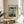 May include: Framed Claude Monet exhibition poster featuring Woman with a Parasol painting, displayed on light wood round side table next to beige upholstered chair. Poster shows impressionist artwork of woman in flowing dress holding green parasol with child in grassy field under cloudy blue sky. Natural lighting creates soft shadows across neutral-toned room with white curtains and plant leaves visible in corner.