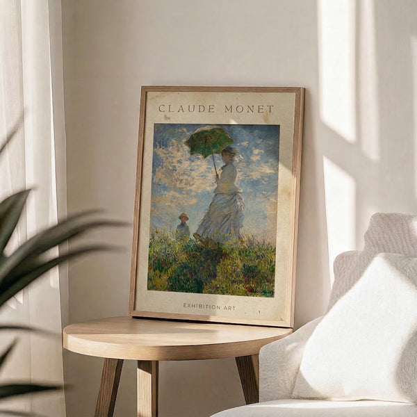 May include: Framed Claude Monet exhibition poster featuring Woman with a Parasol painting, displayed on light wood round side table next to beige upholstered chair. Poster shows impressionist artwork of woman in flowing dress holding green parasol with child in grassy field under cloudy blue sky. Natural lighting creates soft shadows across neutral-toned room with white curtains and plant leaves visible in corner.