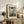 May include: Framed Claude Monet exhibition poster showing woman with green parasol and child in grassy field under cloudy blue sky, displayed on rustic wooden sideboard alongside fern plant, ceramic vase, and stacked books in neutral-toned living room setting