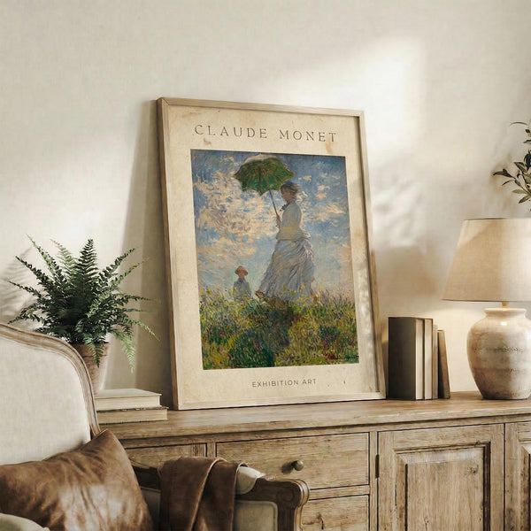 May include: Framed Claude Monet exhibition poster showing woman with green parasol and child in grassy field under cloudy blue sky, displayed on rustic wooden sideboard alongside fern plant, ceramic vase, and stacked books in neutral-toned living room setting