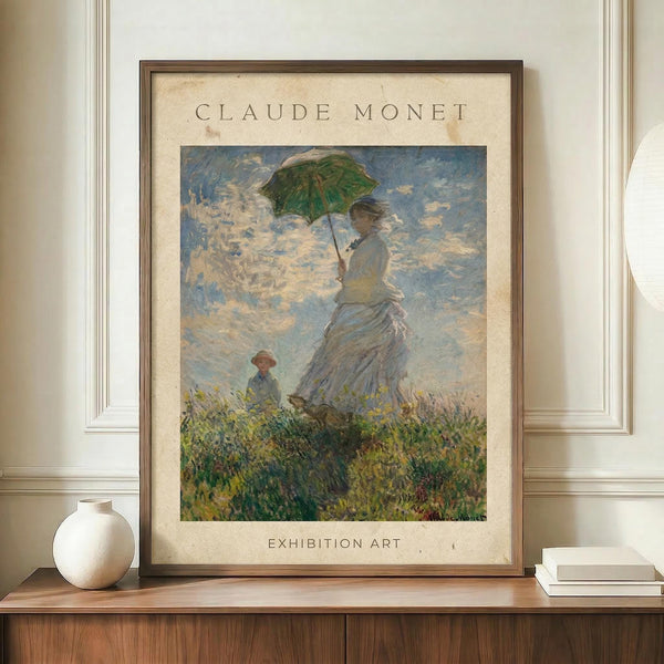 May include: Framed Claude Monet exhibition poster featuring Woman with a Parasol painting, showing a woman in flowing white dress holding green parasol with small child in wildflower meadow under cloudy sky. Warm wood frame with cream matting displays impressionist artwork on wooden sideboard beside white ceramic vase and stacked books against paneled wall.