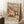 May include: Framed vintage-style beach scene poster leaning against white paneled wall on fluffy white rug. Shows sandy beach with pink and blue umbrellas, sunbathers, and people walking along shoreline. Text reads "COASTAL" at top and "ART POSTER NO. 2" at bottom in navy blue letters on cream background. Light wood frame, displayed next to wooden side table with white coffee cup and saucer, beige throw blanket visible at right edge.