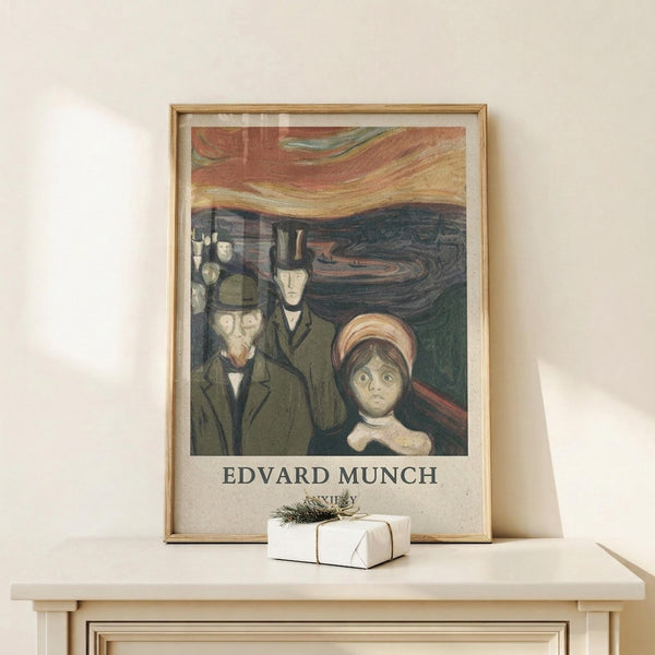 May include: Framed art print featuring Edvard Munch's expressionist painting with figures in dark coats against a swirling orange and red sky, displayed on a light wood console table next to a small white wrapped gift with pine sprig. The poster shows anxious-looking people in muted green and brown tones with dramatic brushstrokes in the background sky.