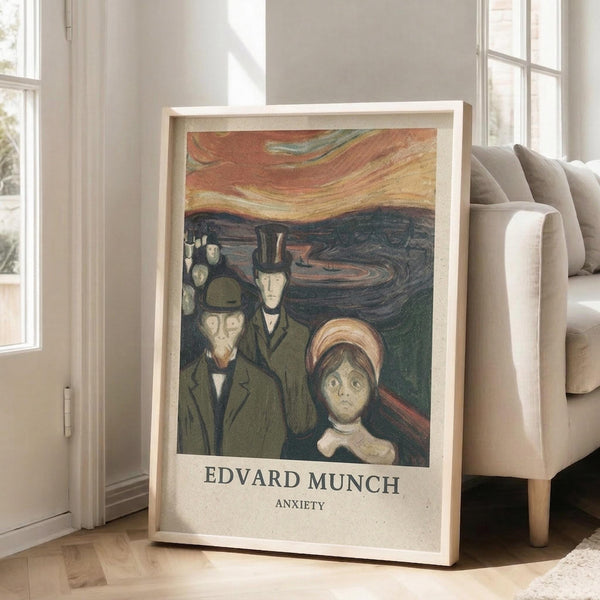 May include: Framed art print leaning against white wall next to beige sofa in bright room with large windows. Print features Edvard Munch's Anxiety painting showing distorted figures in dark coats against swirling orange sky and turbulent landscape. Light wood frame with white matting. Natural lighting creates soft shadows on wooden floor.