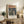 May include: Framed art print displayed on rustic wooden sideboard in neutral-toned room setting. The poster features Munch's expressionist painting showing three figures against a turbulent orange sky and dark landscape. Visible alongside fern plant, cream ceramic vase, stacked books, and table lamp on wood furniture with white upholstered chair in foreground.