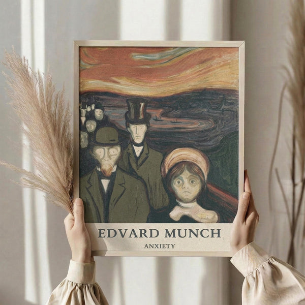May include: Person in cream ruffled sleeve top holding framed art print against white curtained window, with dried wheat stalks visible to the left. The poster features Edvard Munch's expressionist painting showing several figures in dark coats and top hats with a prominent figure in foreground having wide, anxious eyes, set against swirling orange and dark green background. Light wooden frame surrounds the artwork.