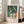 May include: Framed art print leaning against white wall next to beige sofa in bright room with large windows. Print shows Edvard Munch's expressionist painting of nude figures swimming and diving in teal-green water, with yellow and orange skin tones. Text below reads "EDVARD MUNCH BOYS BATHING" on cream background. Light wood frame, soft natural lighting creates shadows on wooden floor.