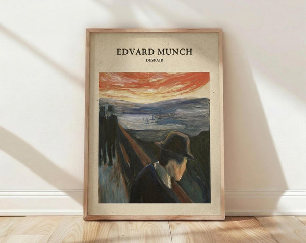 May include: Framed art print featuring Edvard Munch's "Despair" painting showing a figure in dark clothing and hat against a dramatic landscape with orange-red streaked sky and rolling hills. The poster has vintage beige background with black text at top. Natural wood frame sits on light wooden floor against white wall with soft window light creating shadows.