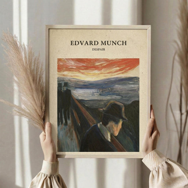 May include: Framed art print featuring Munch's expressionist painting of a figure in dark clothing and hat on a winding road, with dramatic orange-red sky and moody landscape. Person in cream sweater holds the beige-framed poster alongside dried wheat stalks against white curtained window background.