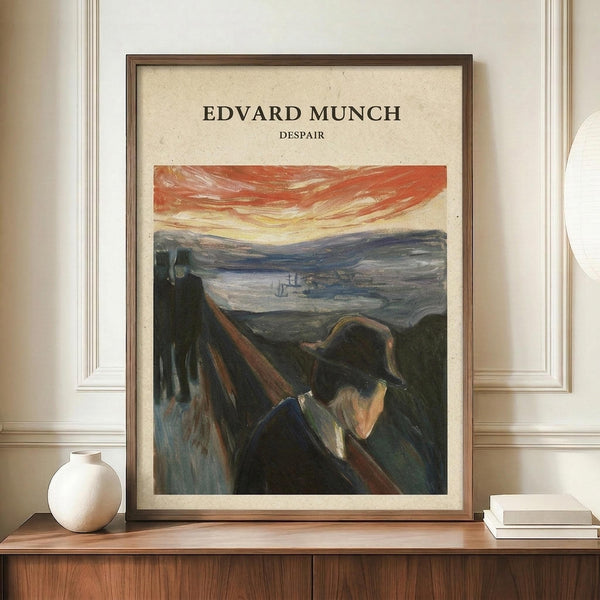 May include: Framed art print displaying an expressionist painting of a solitary figure in dark clothing against a dramatic landscape with swirling orange and red sky. The artwork shows bold brushstrokes and moody colors typical of early 20th century expressionism. The print is mounted in a thin brown frame and positioned on a wooden sideboard alongside a small white ceramic vase and stacked books in a modern interior with white paneled walls.