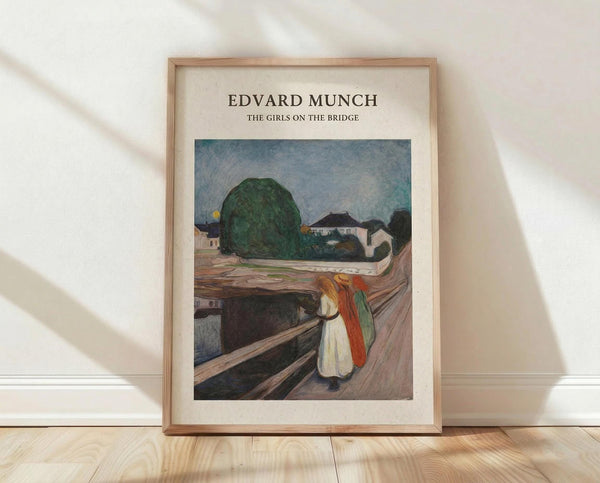 May include: Framed art print of Edvard Munch's "The Girls on the Bridge" showing three figures in white and colorful clothing standing on a wooden bridge overlooking a landscape with green trees and buildings. The reproduction features muted colors and expressionist brushwork. Light wood frame with cream matting, positioned on wooden floor against white wall with natural sunlight casting diagonal shadows across the scene.