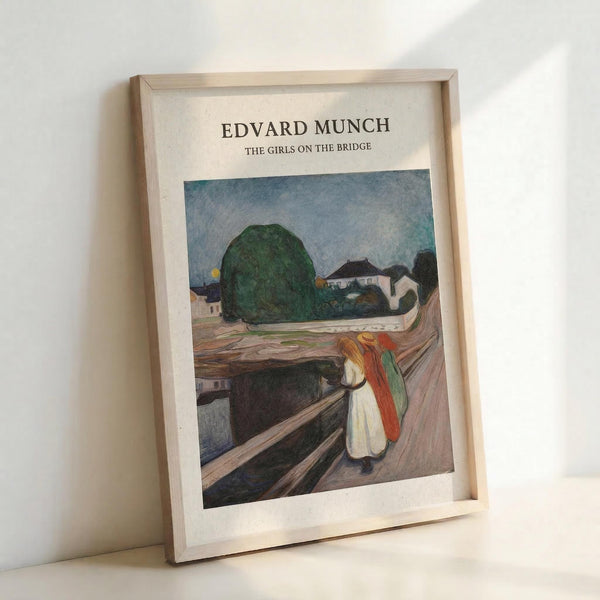 May include: Framed art print in light wood frame showing Edvard Munch's expressionist painting of three figures in colorful clothing standing on a wooden bridge. The artwork features muted colors with a large green tree, small houses, and cloudy sky in the background. The frame leans against a white wall casting soft shadows.