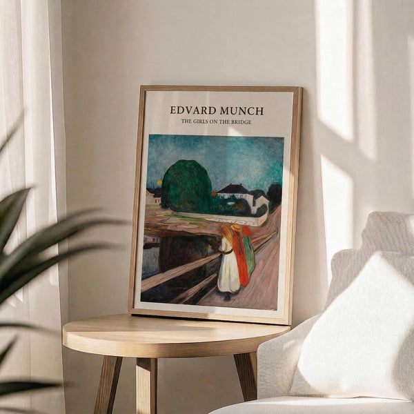 May include: Framed art print of Edvard Munch's "The Girls on the Bridge" displayed on a light wood round side table next to a beige upholstered chair. The poster shows three figures in white and red dresses standing on wooden bridge planks, with green trees and blue-teal sky in background. Thin natural wood frame, soft window lighting creates shadows on white wall, green plant leaves visible in corner.