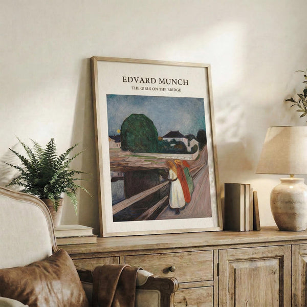 May include: Framed art print of Edvard Munch's expressionist painting showing figures on a wooden bridge with a large green tree and buildings in the background, displayed on a rustic wooden sideboard alongside books, a potted fern, ceramic vase, and brown leather chair in a neutral-toned living room setting.
