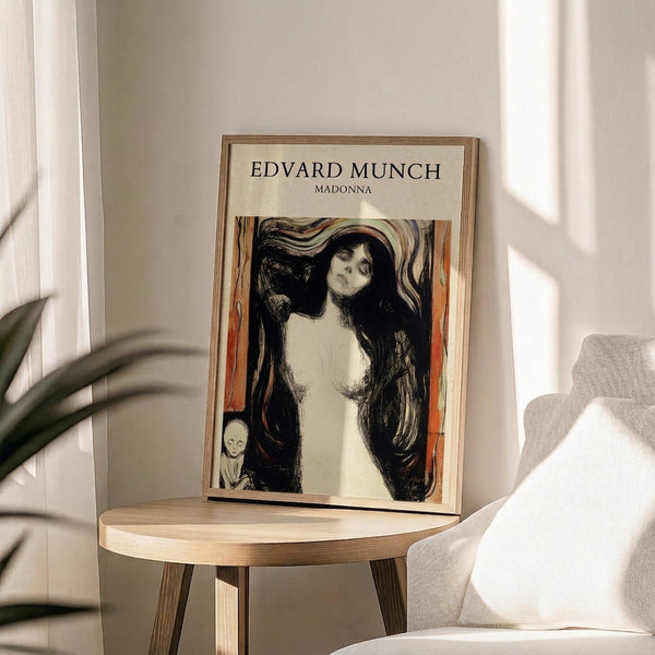 May include: Framed art print featuring Edvard Munch's Madonna artwork showing a woman with long dark hair and pale skin against flowing orange and black brushstrokes, with a small skeletal figure in the corner. The print sits on a light wood round side table next to a beige upholstered chair, with plant leaves visible and natural sunlight casting shadows on white walls.