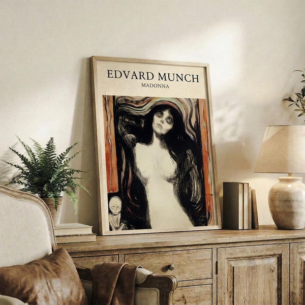 May include: Framed art print featuring Edvard Munch's Madonna artwork showing a woman with long dark hair and pale skin against swirling dark background with orange-red vertical elements and small skull detail. Light wood frame displayed on rustic wooden sideboard alongside fern plant, ceramic vase, books, and table lamp in neutral-toned living room setting.