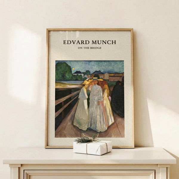 May include: Framed art print displaying Edvard Munch's expressionist painting of three figures in flowing white and yellow dresses standing on a wooden bridge. The artwork features bold brushstrokes with a landscape background of green trees, buildings, and blue sky. Print has cream matting in natural wood frame, displayed on light wooden surface alongside small white gift box with greenery sprig.