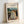 May include: Framed art print leaning against white wall showing Edvard Munch's painting of four figures in flowing dresses standing on wooden bridge planks. The expressionist artwork features women in white and yellow gowns against moody blue-green landscape with buildings and moon visible in background. Simple light wood frame surrounds the reproduction with "EDVARD MUNCH ON THE BRIDGE" text at top.