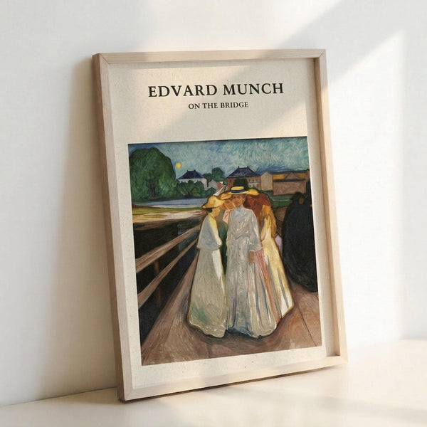May include: Framed art print leaning against white wall showing Edvard Munch's painting of four figures in flowing dresses standing on wooden bridge planks. The expressionist artwork features women in white and yellow gowns against moody blue-green landscape with buildings and moon visible in background. Simple light wood frame surrounds the reproduction with "EDVARD MUNCH ON THE BRIDGE" text at top.