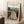 May include: Framed poster print leaning against white wall showing Edvard Munch painting of three figures in flowing white and yellow dresses standing on wooden bridge. Expressionist style artwork with muted colors, rural buildings and trees in background. Light wood frame, displayed next to coffee cup on wooden block and beige textured rug in minimalist room setting.