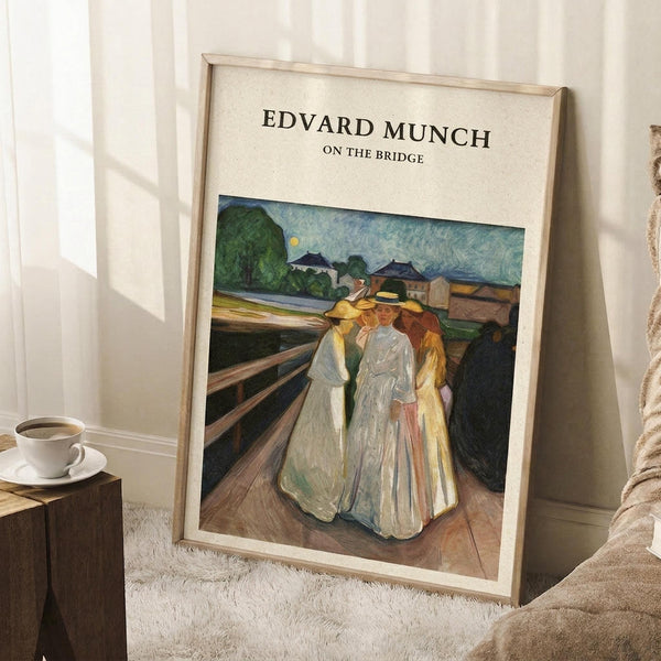 May include: Framed poster print leaning against white wall showing Edvard Munch painting of three figures in flowing white and yellow dresses standing on wooden bridge. Expressionist style artwork with muted colors, rural buildings and trees in background. Light wood frame, displayed next to coffee cup on wooden block and beige textured rug in minimalist room setting.