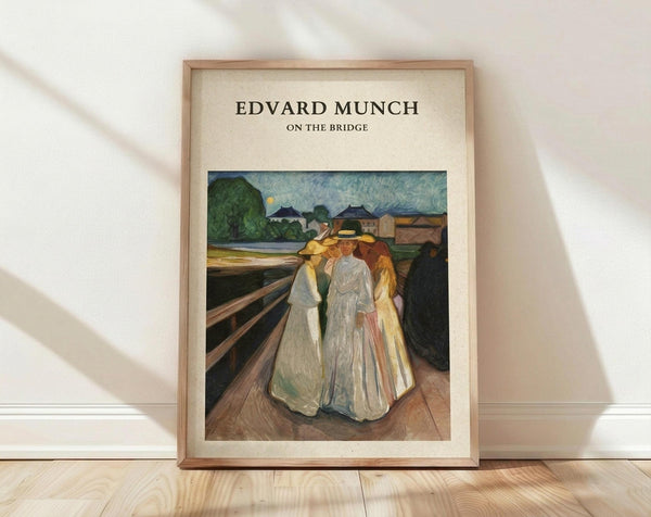 May include: Framed art print leaning against white wall on light wooden floor. The poster features Edvard Munch's painting of three women in white dresses standing together on a wooden bridge. The artwork shows an evening scene with buildings and trees in the background under a pale sky. The print has cream-colored matting with "EDVARD MUNCH ON THE BRIDGE" text above the image, all contained within a thin light wood frame.